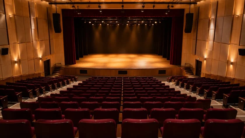 Plateia vazia de um teatro com poltronas vermelhas e palco iluminado